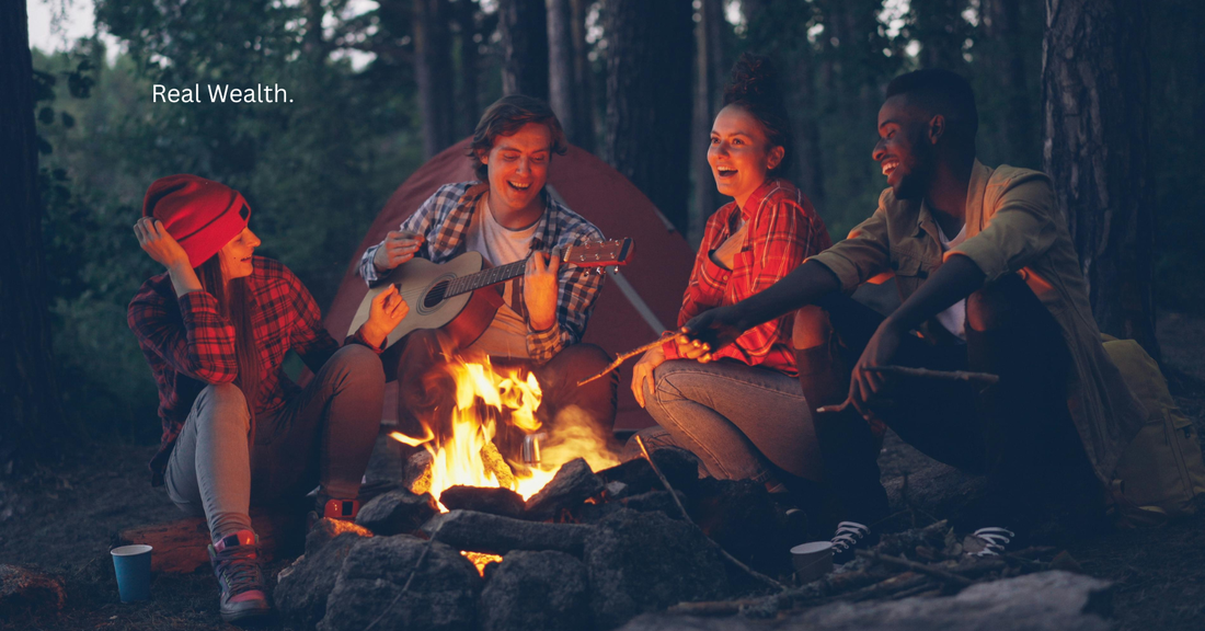 Friends gathered around a campfire in the woods at night, sharing music, laughter, and connection as a reflection of real wealth, presence, and intentional living.